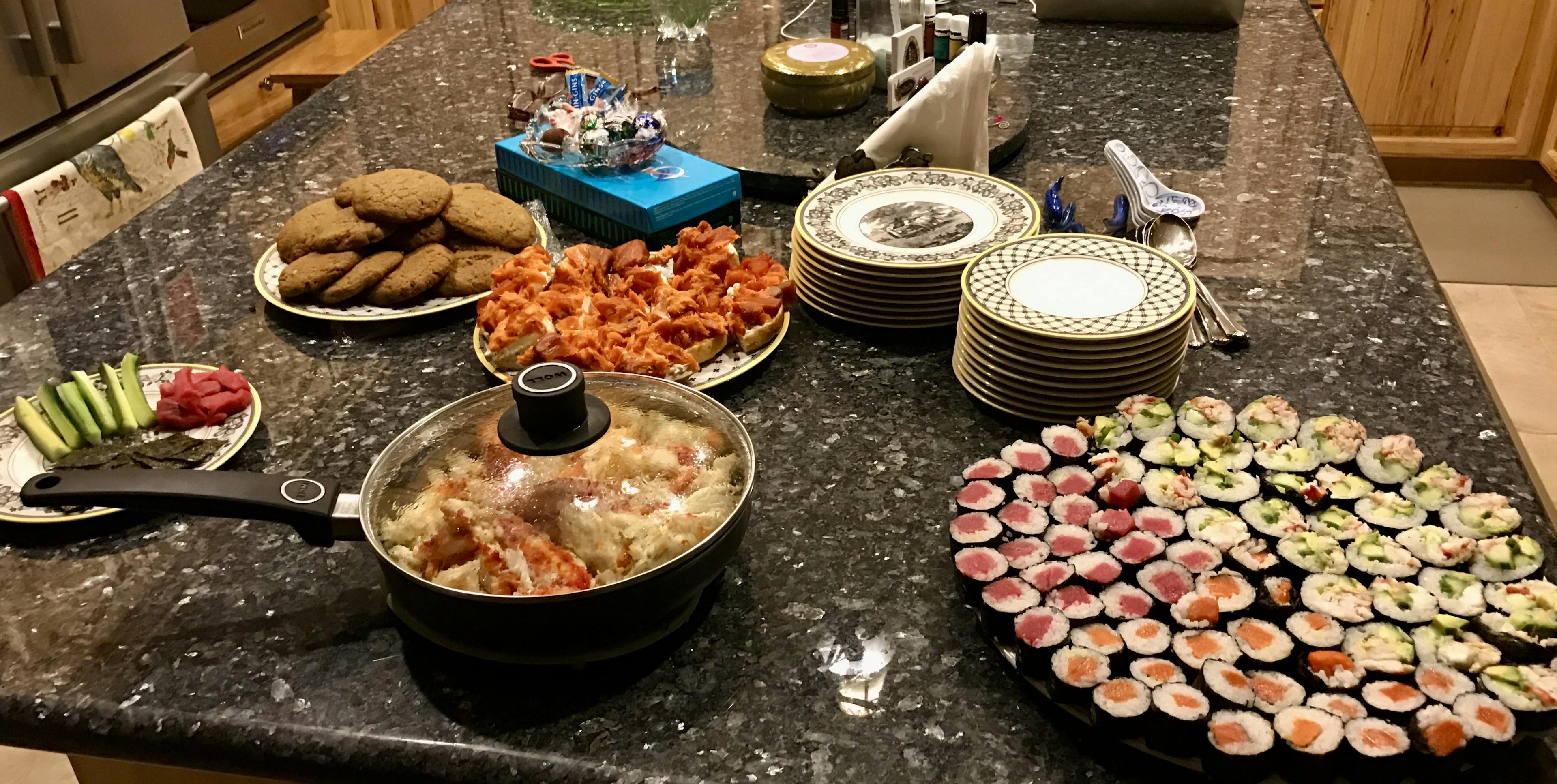 Kitchen table with populated with a large platter maki rolls, cookies, and other assortments 