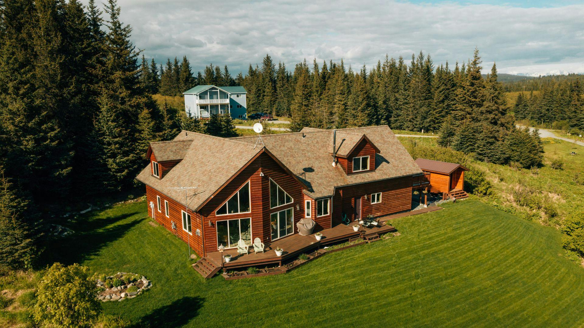 Aerial view of a large log cabin home surrounded by trees with floor to ceiling picture windows, back deck overlooking expansive lawn