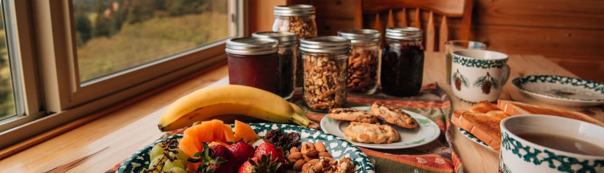 Table by a window full of fruit, jars of jams and granola and mugs of coffee