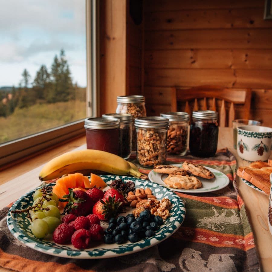 Breakfast table by a large window with plates of fruit and pastries, jars of jams and granola and mugs of coffee